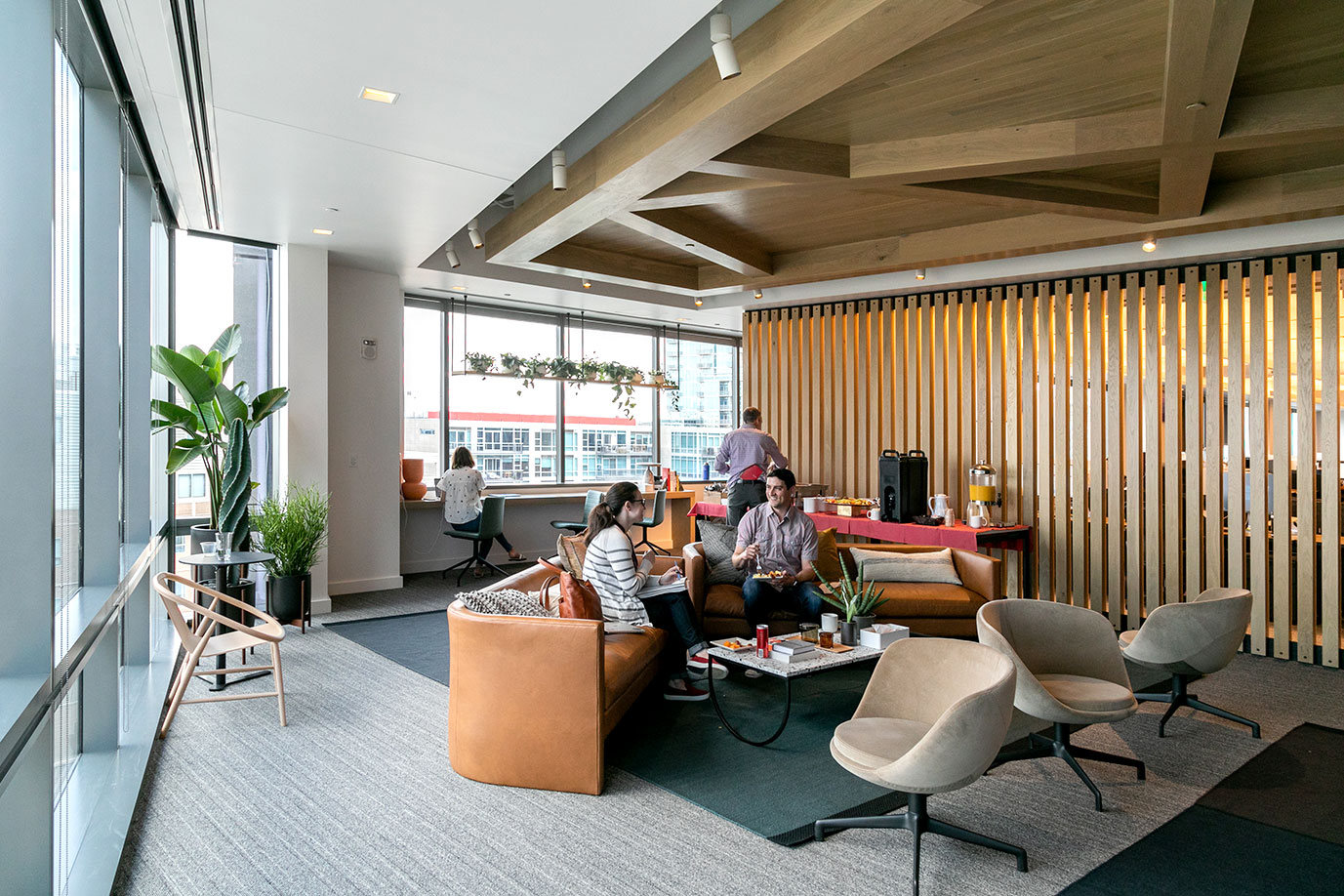 An open office area space featuring big windows, wooden architectural details in a West Coast style of design, and two employees reposing on a plush sofa.